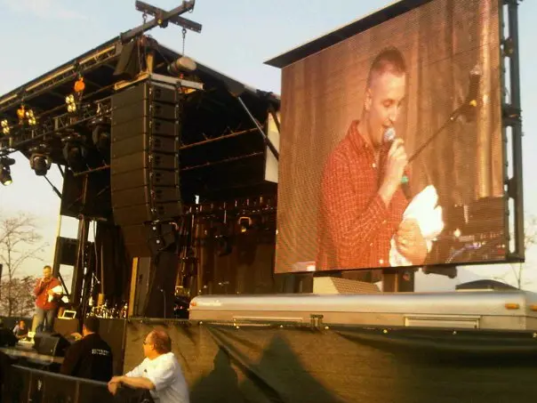 Rob Daniels on stage at a concert, shown on the jumbotron addressing the crowd at golden hour
